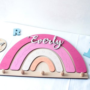 May include: A wooden rainbow-shaped wall decor in shades of pink, peach, and white, with the name "Everly" in cursive. It features decorative cutouts and wooden pegs for hanging items. Additional wooden letters and a round sign are also present.