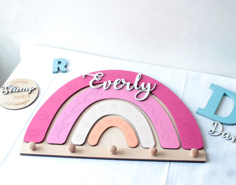 May include: A wooden rainbow-shaped wall decor in shades of pink, peach, and white, with the name "Everly" in cursive. It features decorative cutouts and wooden pegs for hanging items. Additional wooden letters and a round sign are also present.