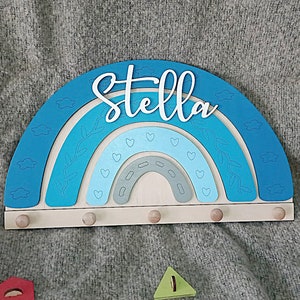 May include: A wooden rainbow-shaped wall decor with the name "Stella" in white script. The rainbow features shades of blue and gray, with heart and leaf details. It has five small wooden pegs for hanging items.