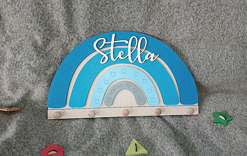 May include: A wooden rainbow-shaped wall decor with the name "Stella" in white script. The rainbow features shades of blue and gray, with heart and leaf details. It has five small wooden pegs for hanging items.