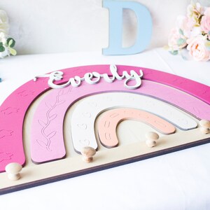 May include: A wooden rainbow-shaped wall decor in shades of pink, with the name "Everly" in white script. The design includes decorative cutouts and wooden knobs for hanging items. The piece is approximately 12 inches wide.