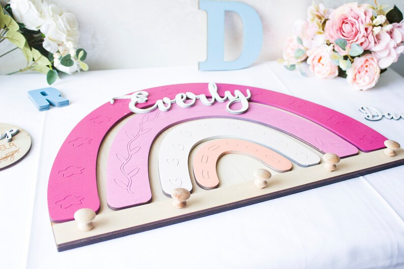 May include: A wooden rainbow-shaped wall decor in shades of pink, with the name "Everly" in white script. The design includes decorative cutouts and wooden knobs for hanging items. The piece is approximately 12 inches wide.