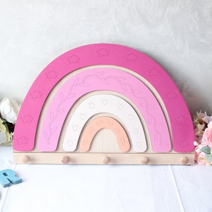 May include: A wooden rainbow-shaped wall organizer in shades of pink, peach, and white. The design features cloud cutouts, leaf and heart details, and six wooden pegs for hanging items. A decorative piece for a nursery or child's room.