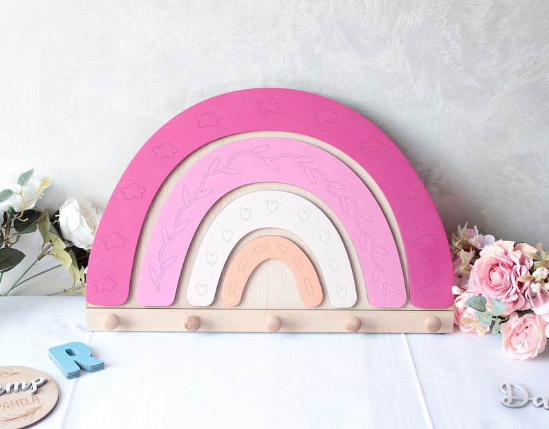 May include: A wooden rainbow-shaped wall organizer in shades of pink, peach, and white. The design features cloud cutouts, leaf and heart details, and six wooden pegs for hanging items. A decorative piece for a nursery or child's room.