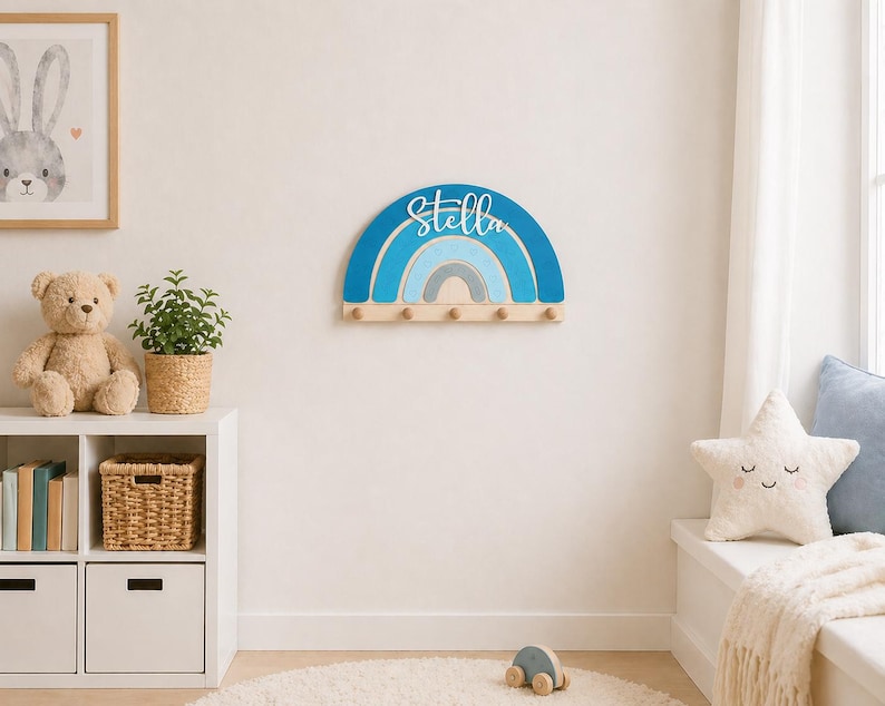 May include: A wooden rainbow-shaped wall hook with the name "Stella" in blue script. The rainbow features shades of blue and gray. Below the rainbow are several wooden pegs for hanging items. The wall decor is mounted on a white wall.