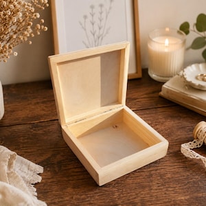 May include: A square, light-colored wooden box with an open hinged lid. The box features a smooth, unfinished natural wood grain texture, a simple square frame construction, and a flat interior base, presented as a standalone storage container.