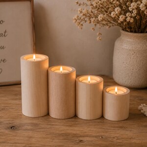 Pode incluir: Um conjunto de quatro castiçais cilíndricos de madeira de cor clara com alturas variadas, cada um contendo uma pequena vela de chá acesa com uma chama visível. Os suportes de madeira têm um acabamento liso com grão natural.