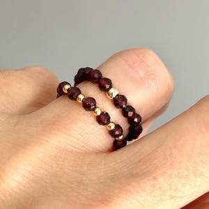 May include: Two stacked rings made of small, faceted, deep red garnet beads interspersed with tiny, polished, round gold-tone metal beads. The rings are worn together on a finger, showcasing the contrast between the dark red stones and gold.