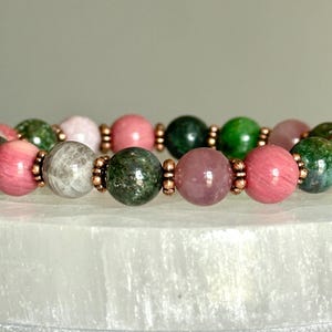 May include: A beaded bracelet featuring alternating round polished stones in shades of pink, dark green, and grey. The beads are separated by small, metallic copper-coloured spacers, creating a repeating pattern throughout the circular jewellery piece.