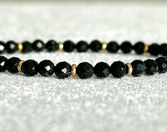 May include: A beaded bracelet featuring small, faceted black beads arranged in a repeating pattern with thin, polished gold-tone metal spacers placed between the beads. The bracelet has a circular shape and a reflective, dark, and metallic finish.