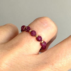 May include: A ring featuring a series of small, faceted, deep magenta-colored beads alternating with tiny, smooth, polished gold-toned spherical beads. The circular band is constructed from these repeating bead elements arranged in a continuous pattern.