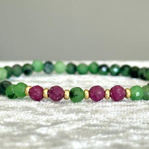 May include: A beaded bracelet featuring small, faceted green gemstone beads interspersed with three larger, round, deep pink ruby beads. The design includes small, polished gold-tone metal spacer beads between the ruby and green stones.