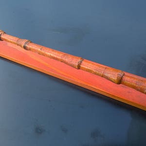 May include: A wooden wand with a reddish-brown finish, mounted on a wooden stand with a red stain. The wand is made of multiple pieces of wood, giving it a segmented appearance.