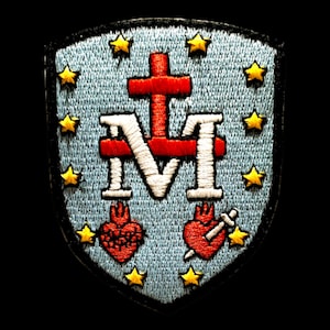 Könnte beinhalten: Ein blauer gestickter Aufnäher mit einem weißen "M" und einem roten Kreuz. Zwei rote Herzen mit Flammen und einem Schwert befinden sich unter dem "M". Der Aufnäher ist von einem Rand aus gelben Sternen umgeben.