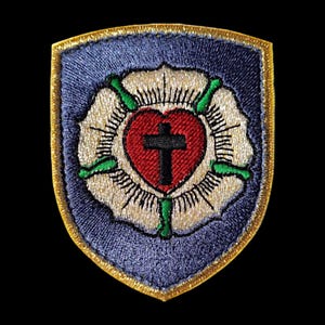 Può includere: Una toppa ricamata blu e oro con una rosa bianca, un cuore rosso e una croce nera. Il cuore è al centro della rosa e la croce è all'interno del cuore.