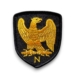 May include: A black embroidered patch featuring a gold eagle and the letter "N" in the centre. The eagle is a symbol of France.