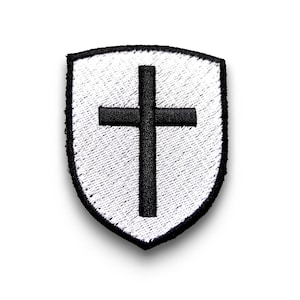 Peut inclure: Un écusson brodé blanc en forme de bouclier avec une croix noire au centre.