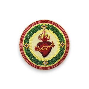 Könnte beinhalten: Runder, gestickter Aufnäher mit einem roten Herz und einer Dornenkrone, gekrönt von einem Kreuz. Das Herz ist von einem grünen und gelben Kranz und einem roten Rand umgeben. Der Aufnäher hat einen cremefarbenen Hintergrund.