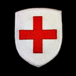 Può includere: Una toppa ricamata bianca a forma di scudo con una croce rossa al centro.