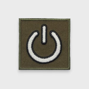Peut inclure: Un écusson brodé de forme carrée présentant un fond vert olive avec une icône de symbole d'alimentation blanche centrée au milieu. Le symbole d'alimentation est souligné par un fil noir, et l'écusson possède une bordure cousue vert olive.