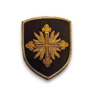 Op de afbeelding: Een zwarte geborduurde patch met een gouden kruis omringd door een zonnestralenontwerp. De patch heeft de vorm van een schild.