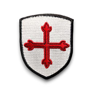 Peut inclure: Un écusson brodé blanc avec une croix rouge au centre. La croix a un contour noir et est entourée d'une bordure noire. L'écusson a la forme d'un bouclier.