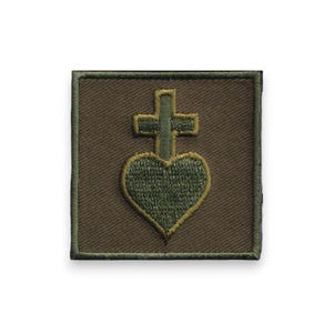 Può includere: Una toppa quadrata verde oliva con un bordo cucito. La toppa presenta una forma di cuore verde con un contorno dorato, sormontata da una croce verde. Il disegno è centrato sulla toppa.