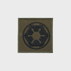Op de afbeelding: Een vierkante olijfgroene geborduurde patch met een donker marineblauw cirkelvormig embleem in het midden. Het embleem bestaat uit een gestileerd tandwielontwerp met zes spaken binnen een cirkelvormige rand, alles uitgevoerd in dicht stikwerk.