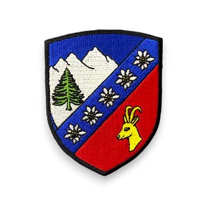 Könnte beinhalten: Gestickter Aufnäher in Schildform. Die obere Hälfte ist blau mit weißen Bergen und einer grünen Kiefer. Die rechte Hälfte ist rot mit einer gelben Ziege. Weiße Blumen befinden sich auf einem blauen Diagonalkreuz.