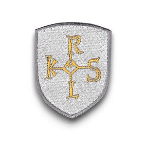 Op de afbeelding: Een zilverkleurige schildvormige patch met een gouden embleem met de letters K, R, S en L. De patch heeft een getextureerd oppervlak en een grijze rand. Het embleem is een gestileerd kruis.