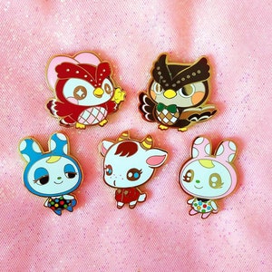 Könnte beinhalten: Fünf Emaille-Pins mit Cartoon-Tierfiguren. Die Pins sind goldfarben und zeigen eine rote Eule, ein blaues Kaninchen, ein rosa Kaninchen, eine braune und weiße Kuh und einen roten Vogel mit weißen Punkten.