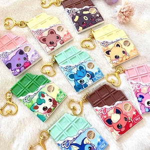 Puede incluir: Una colección de llaveros coloridos con forma de barra de chocolate. Cada llavero presenta un diseño de personaje de dibujos animados diferente y está unido a un cierre dorado en forma de corazón. Los llaveros son de varios colores, incluyendo rosa, verde, amarillo y marrón.