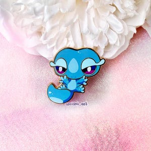 May include: A blue enamel pin featuring a stylised cartoon creature with large eyes, purple pupils, and a long tail. The design includes gold-coloured metal outlines and the text weishi.art printed in small, dark letters near the bottom edge.