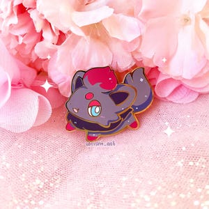 Puede incluir: Un pin con un personaje de dibujos animados con un cuerpo gris, detalles rojos y morados y un contorno dorado. El personaje tiene ojos grandes y una lengua pequeña. El pin está sobre un fondo rosa con flores rosas.