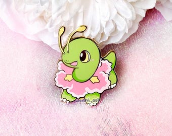 May include: A green enamel pin featuring a cartoon creature with two antennae, a smiling face, and a ruffled pink tutu-like skirt around its midsection. The character has small yellow accents on its sides and is outlined in gold metal.