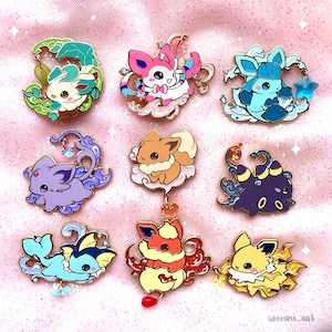 Könnte beinhalten: Neun Emaille-Pins mit verschiedenen Pokémon-Charakteren. Die Pins sind goldfarben und haben eine Vielzahl von Farben und Designs. Jeder Pin hat einen kleinen Anhänger, der daran hängt.