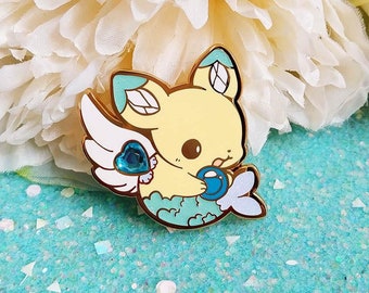 May include: A gold-colored enamel pin featuring a cartoon yellow mermaid with blue wings and a blue heart. The mermaid is holding a blue pearl in its hand.