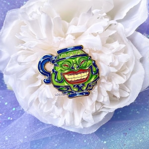 Könnte beinhalten: Ein grün-blauer Emaille-Pin in Form einer lächelnden Teekanne mit rotem Mund und gelben Zähnen. Der Pin hat eine goldene Umrandung und ist auf einer weißen Blume platziert.