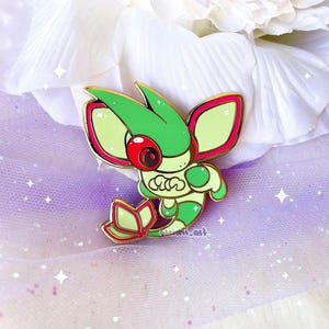 Puede incluir: Un pin de esmalte verde y rojo que representa una criatura estilizada con ojos grandes y un contorno dorado. El pin tiene un borde dorado y está sobre un fondo suave y texturizado. El pin mide aproximadamente 4 cm de altura.