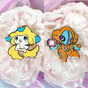 May include: Two enamel pins featuring cartoon characters. One is yellow with blue accents, the other is orange with blue and purple accents. Both pins have a shiny, metallic finish and are set against a backdrop of a large, white flower.