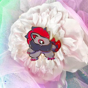 May include: A colorful enamel pin featuring a cartoon-style creature with red, gray, and black accents. The pin is set against a backdrop of a large, white flower and pink fabric.