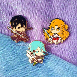 Könnte beinhalten: Drei Emaille-Pins mit Charakteren aus dem Anime Sword Art Online. Die Pins sind goldfarben und zeigen Kirito, Asuna und Leafa. Die Pins befinden sich auf einem glitzernden lila und blauen Hintergrund.