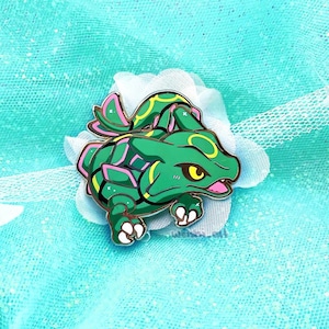 Könnte beinhalten: Eine grüne und rosafarbene Emaille-Anstecknadel mit einer Cartoon-Darstellung eines Pokemon-Charakters. Der Charakter hat einen grünen Körper mit rosa Akzenten und gelben Augen. Die Anstecknadel befindet sich auf einem blau-weißen Hintergrund.