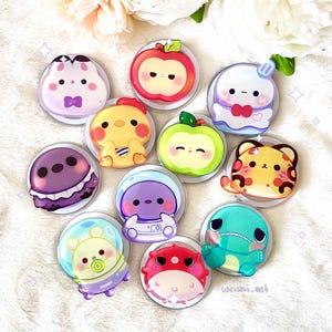 Puede incluir: Una colección de pegatinas redondas y coloridas de personajes de dibujos animados. Las pegatinas presentan varios diseños, como una manzana, un fantasma, un tigre y un astronauta. Las pegatinas están sobre un fondo blanco con un arreglo floral.