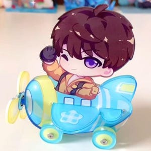May include: A cartoon character with brown hair and purple eyes waves from a blue and yellow airplane toy. The airplane has a propeller and wheels. The character is wearing a yellow and orange outfit.