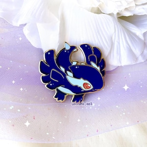 May include: A dark blue enamel pin featuring a stylized dragon design with gold accents. The dragon has white and light blue details, a red eye, and is set against a soft, glittery background. The pin is likely a collectible item.