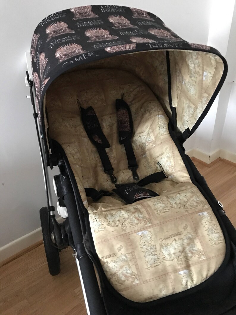 May include: A black stroller with a Game of Thrones themed canopy and seat liner. The canopy is black with a white Game of Thrones logo and the seat liner is beige with a map of Westeros.