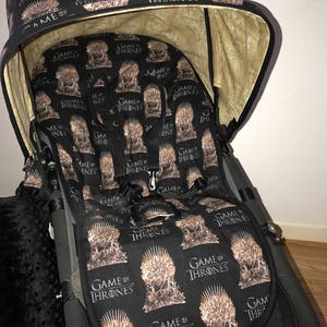 May include: A black stroller with a Game of Thrones themed canopy and seat cover. The canopy is beige on the inside and has the words "Game of Thrones" printed on it. The seat cover is black with a repeating pattern of the Iron Throne and the words "Game of Thrones".