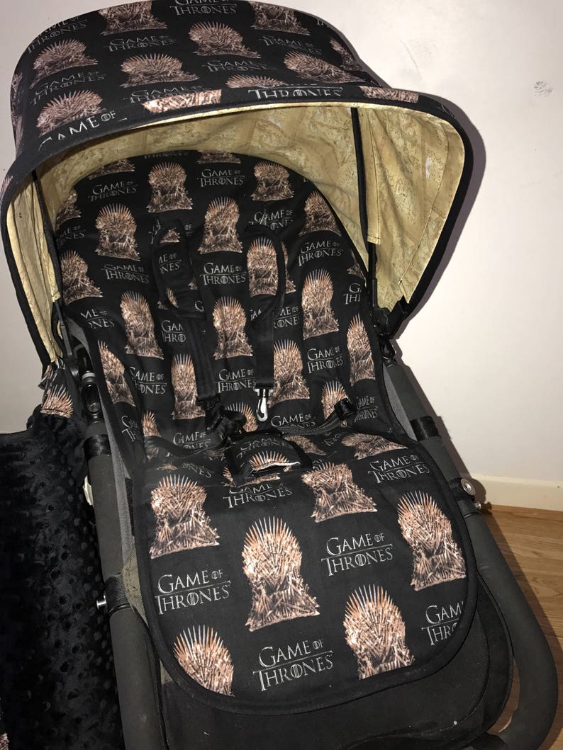 May include: A black stroller with a Game of Thrones themed canopy and seat cover. The canopy is beige on the inside and has the words "Game of Thrones" printed on it. The seat cover is black with a repeating pattern of the Iron Throne and the words "Game of Thrones".