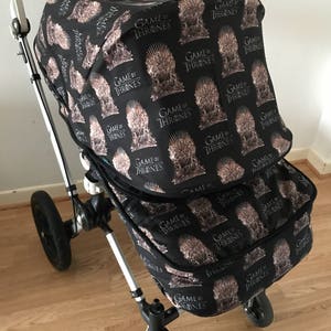 May include: A black stroller with a Game of Thrones themed canopy and storage basket. The canopy and basket are covered in a repeating pattern of the Iron Throne with the words "Game of Thrones" printed on it.